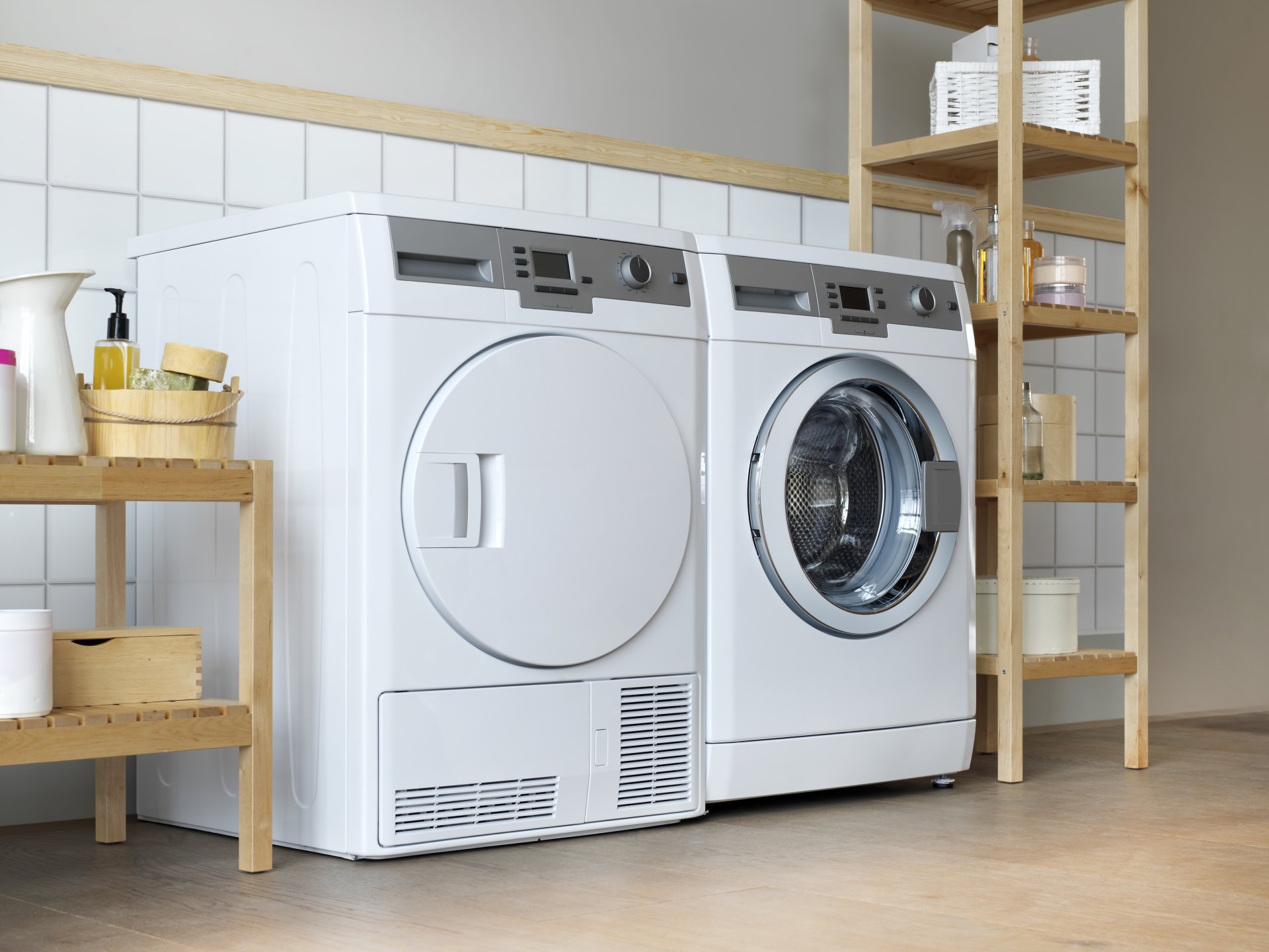 A white front-load washer and dryer in a home laundry room, flanked by wooden shelving holding towels and supplies.