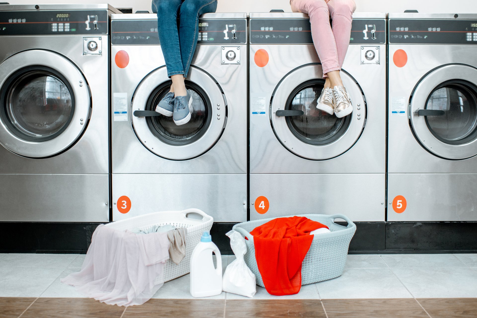 Two people sitting on top of coin-operated washing machines in a shared laundry room, with laundry baskets on the floor below.