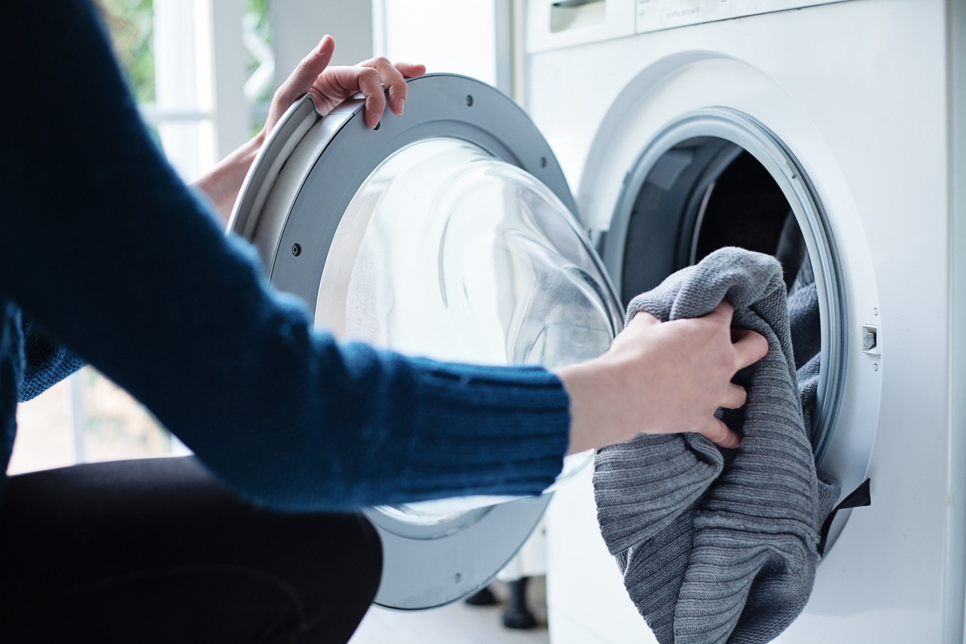 A person loading a gray knit sweater into a front-loading washing machine drum