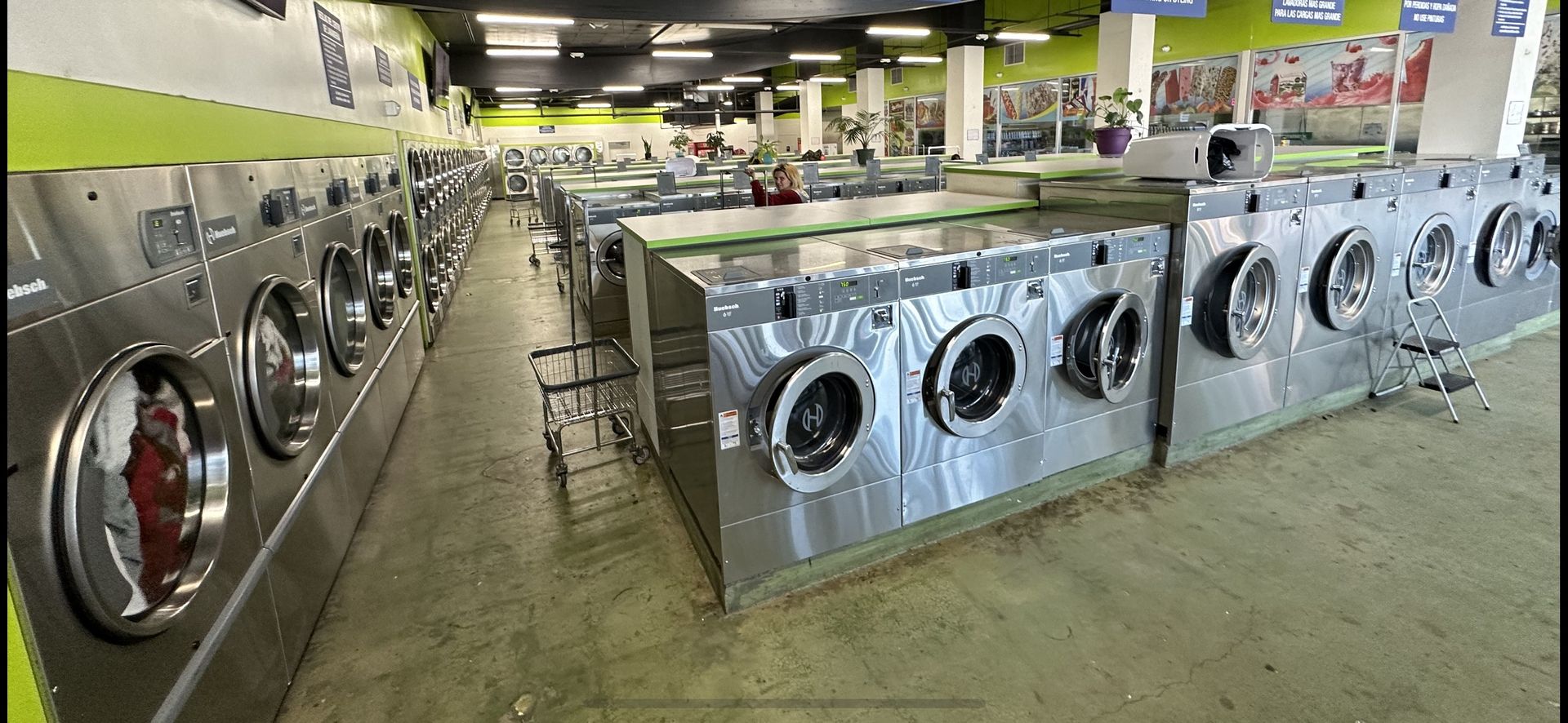 A U-shaped row of stainless steel front-load washers beneath a lime-green wall stripe and black ceiling, with a customer folding laundry at the center island beyond.