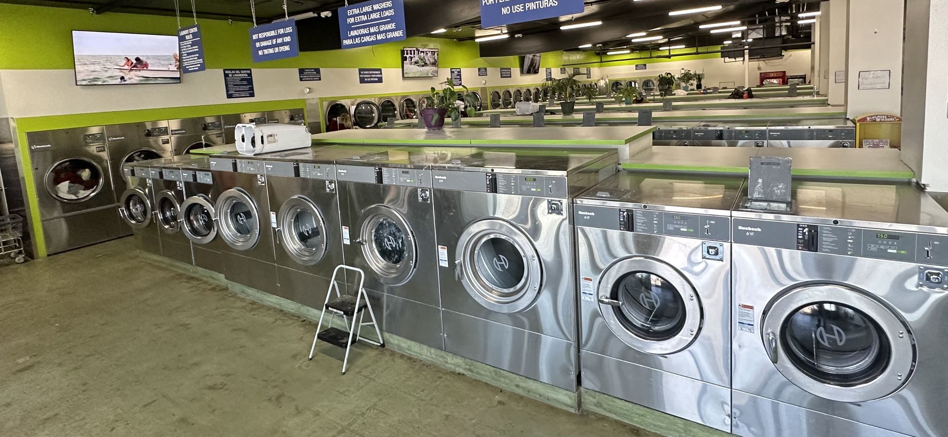 A row of front-load washers along a lime-green wall, with a wall-mounted TV playing overhead and a step stool set out in the aisle.