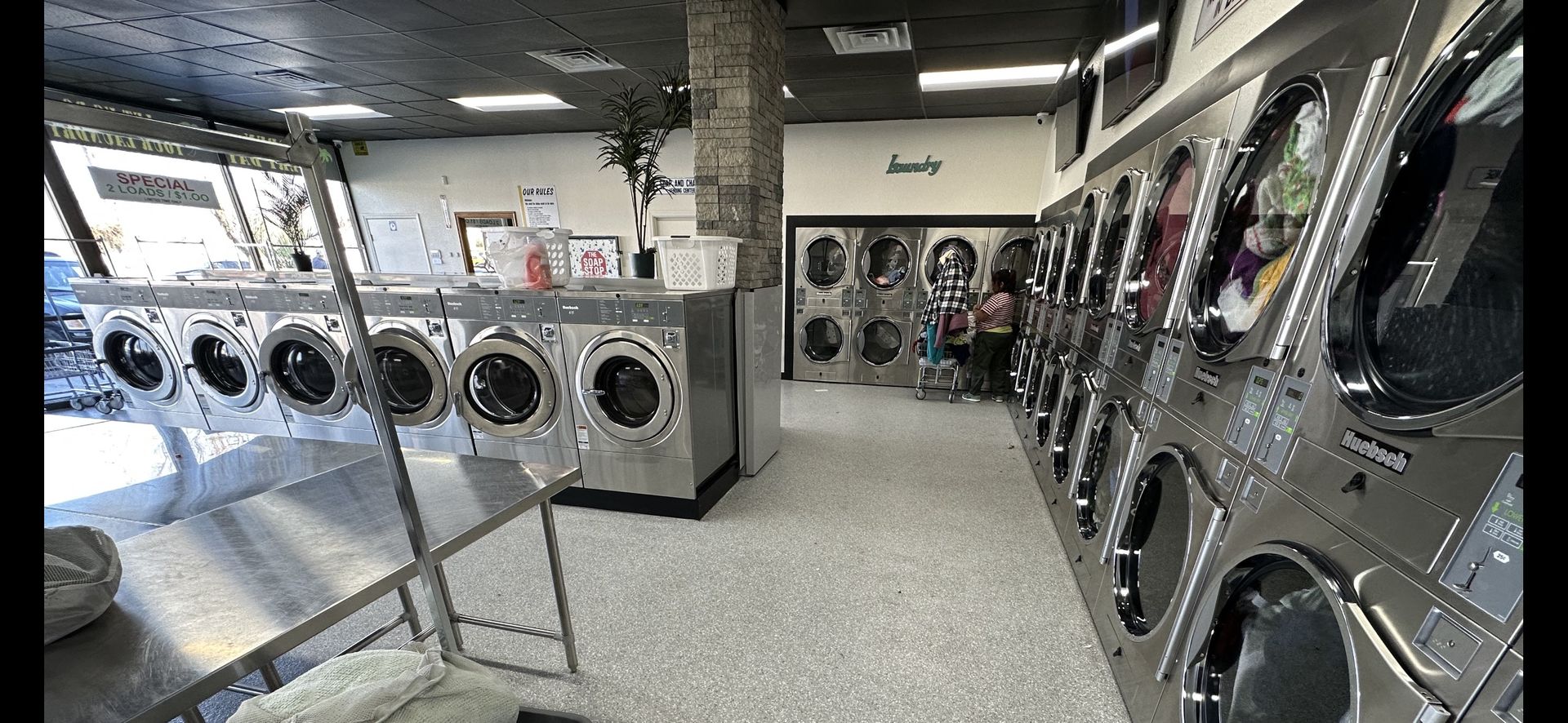 Storefront laundromat interior with a row of front-load washers along the front windows, a stone support column, and a stacked-dryer alcove marked 'Laundry' in the back corner.