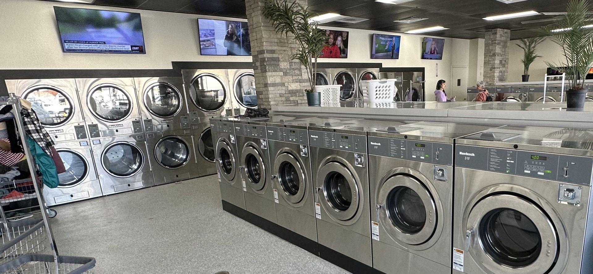 A center aisle between two rows of front-load washers, with ceiling fans overhead, wall-mounted TVs, and a stainless folding table piled with clothes and bags.