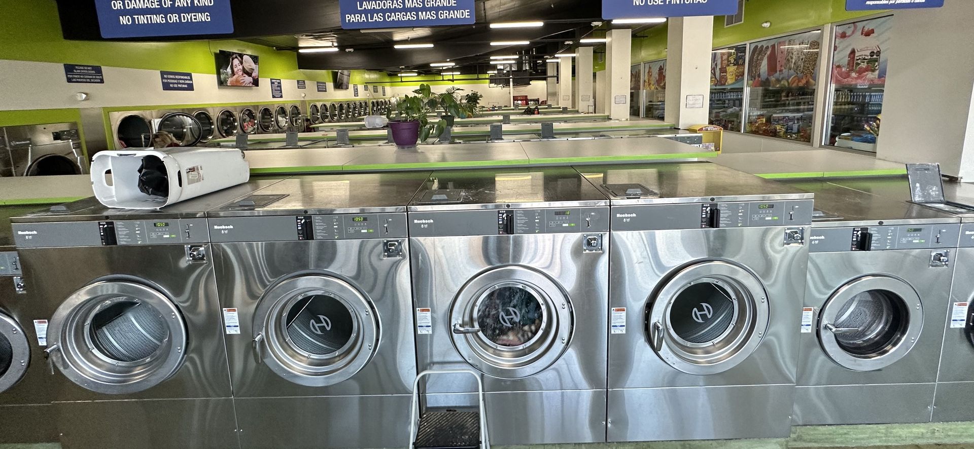 Front-load washers in the foreground beneath bilingual rule signage, with glass-front beverage coolers and a wall-mounted TV visible in the background.