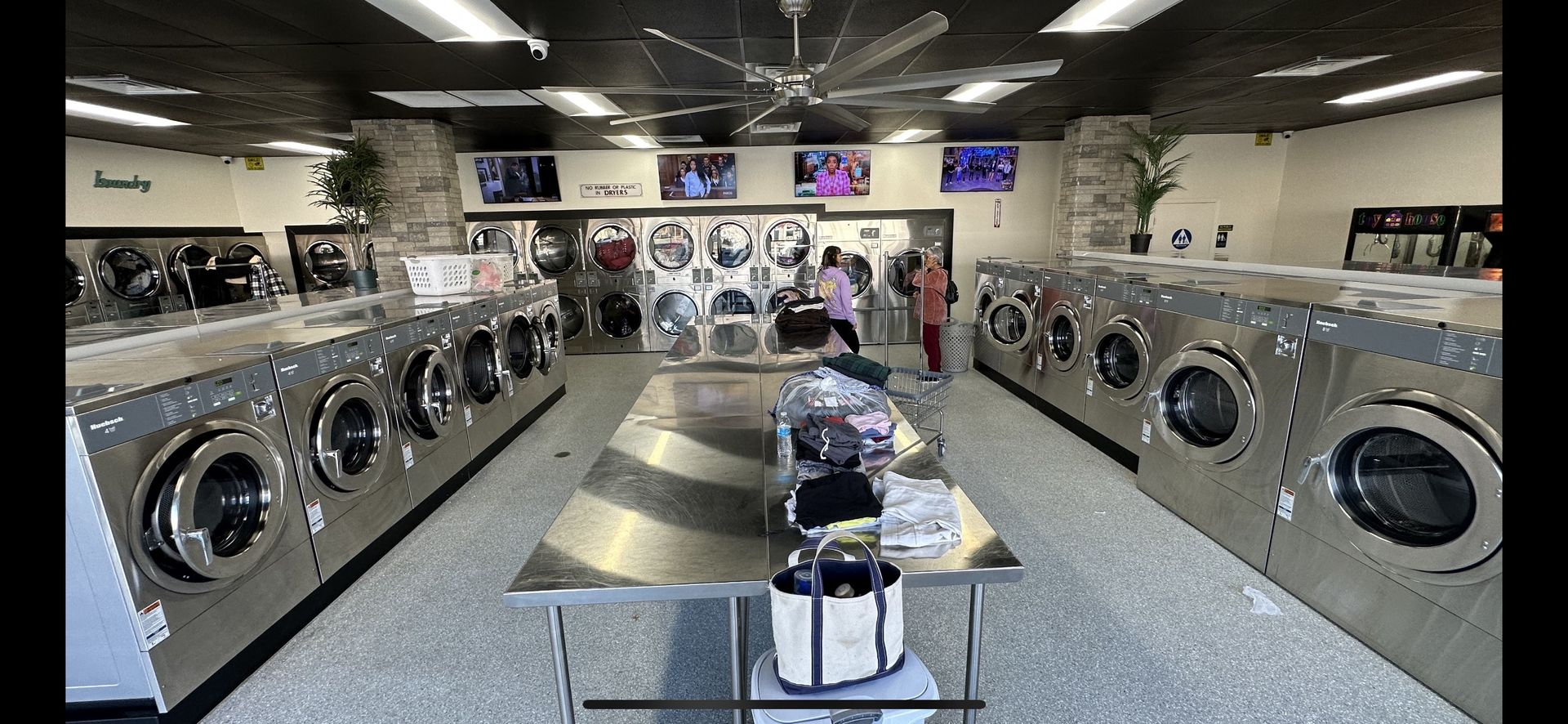 A center aisle of front-load washers under paddle ceiling fans, leading back to a stacked-dryer alcove marked 'Laundry' near the exit door.