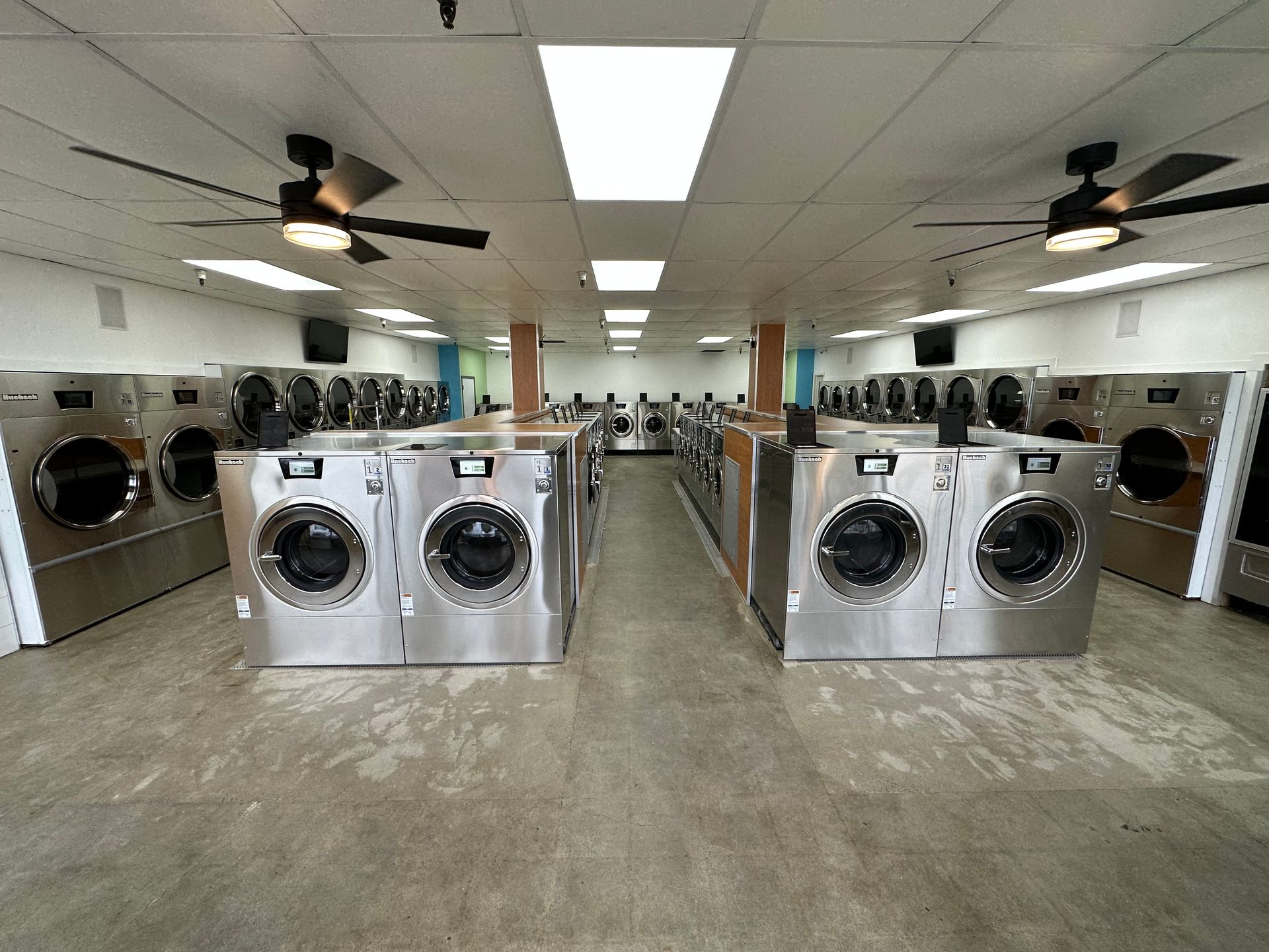 A long aisle of front-load washers with wood-toned end panels and coin-drop pay units mounted at eye level on each machine.