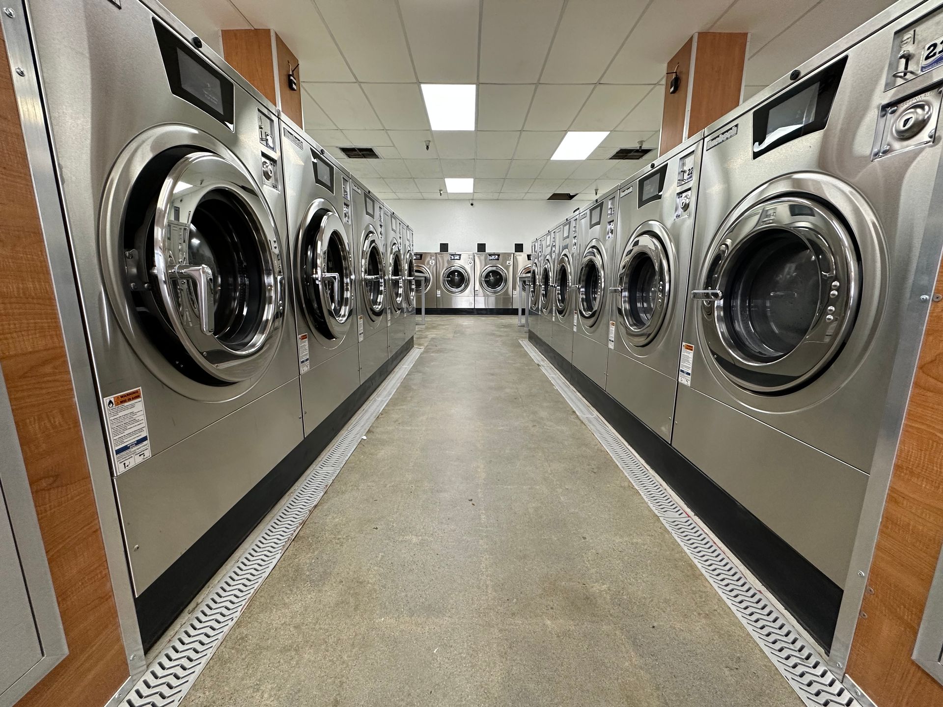 A wide-angle view down a washer aisle with a center island of machines visible between two side rows, lit by overhead ceiling tile fixtures.