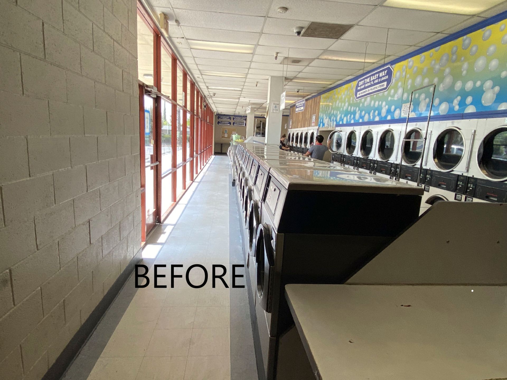 A 'before' documentation photo of the prior laundromat space: a narrow aisle beside red-framed storefront windows, older front-load washers, and a bubble-themed mural on the back wall.