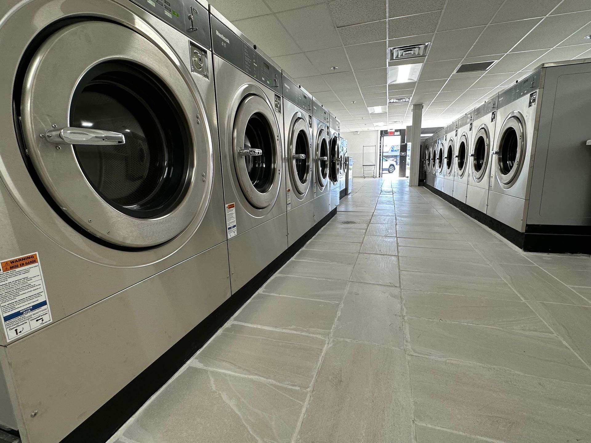 A center aisle of stainless front-load washers, with a neighboring storefront visible through an open door at the far end of the room.