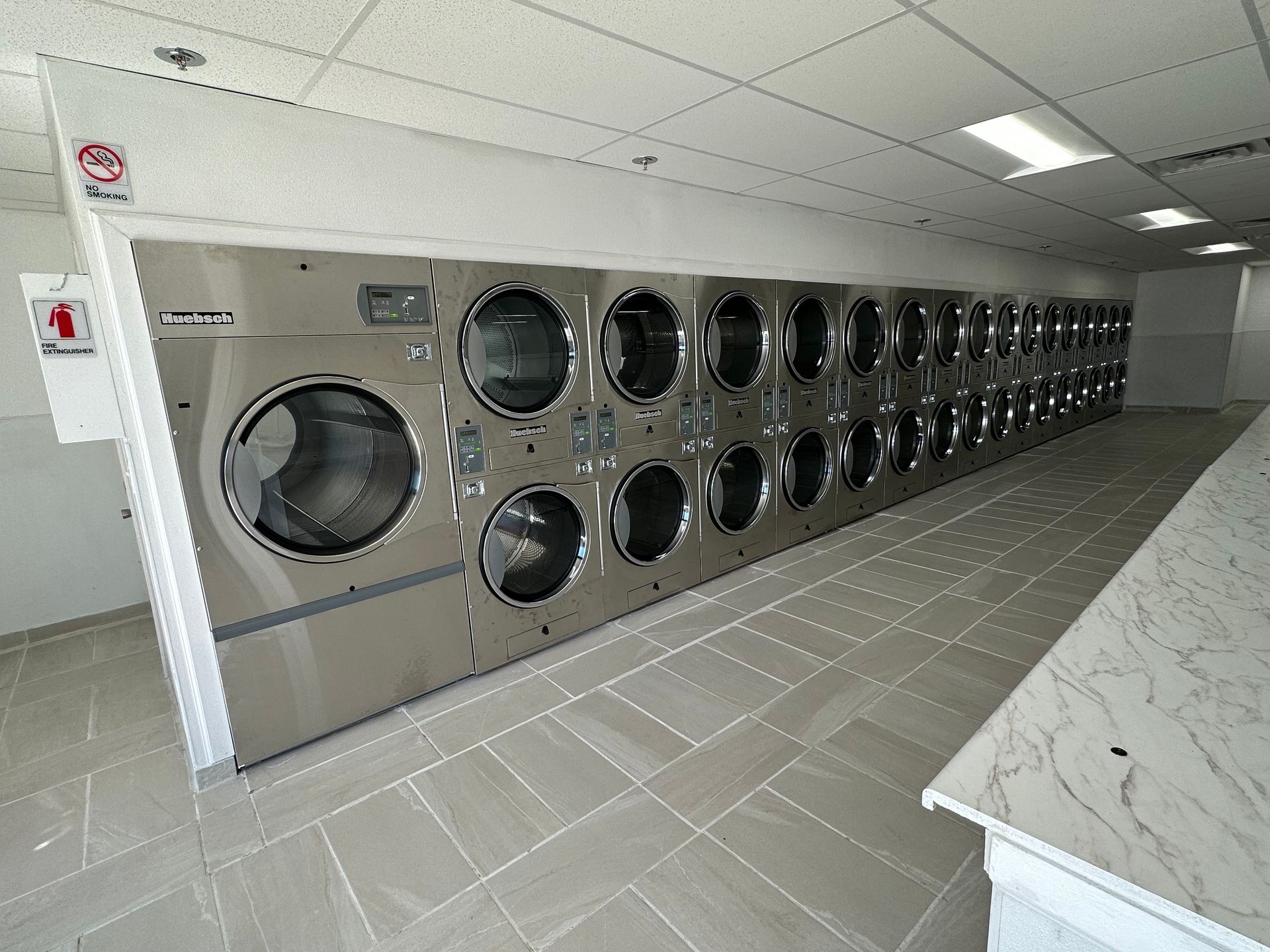 A wall of stacked double dryers beside a marble-look folding counter, with fire extinguisher and no-smoking signage mounted at the entrance.