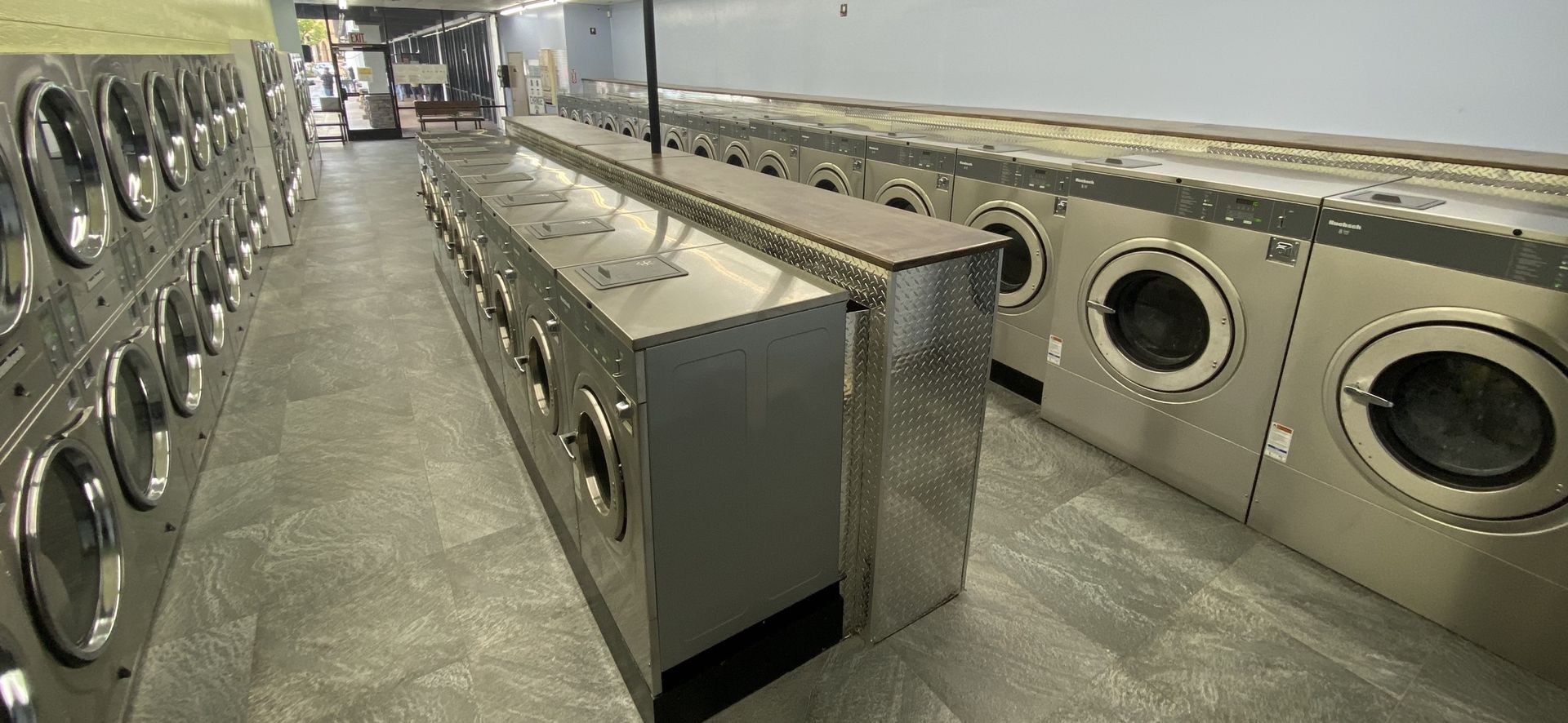 A center aisle of front-load washers with a diamond-plate end panel and a wood-topped folding counter running along the row.