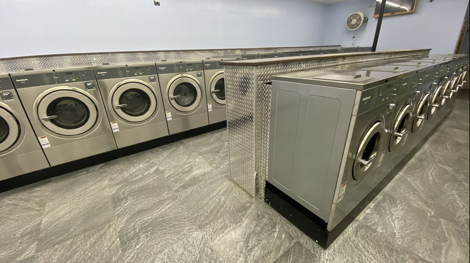 A corner view of a diamond-plate end panel and wood-topped folding counter, with a ceiling fan turning overhead and washers lined up beyond.