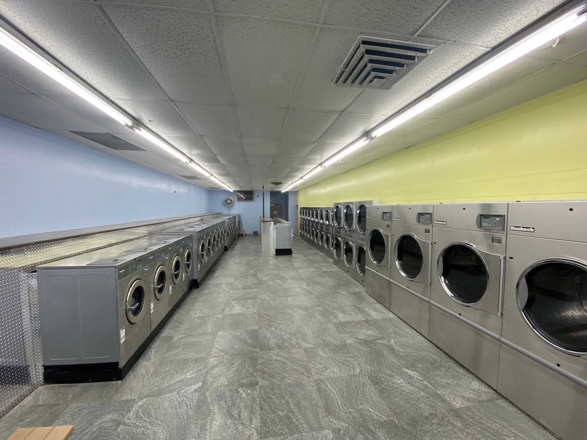 A washer-and-dryer room painted light blue and yellow-green, with a wall-mounted fan and a wood bench along the near wall.