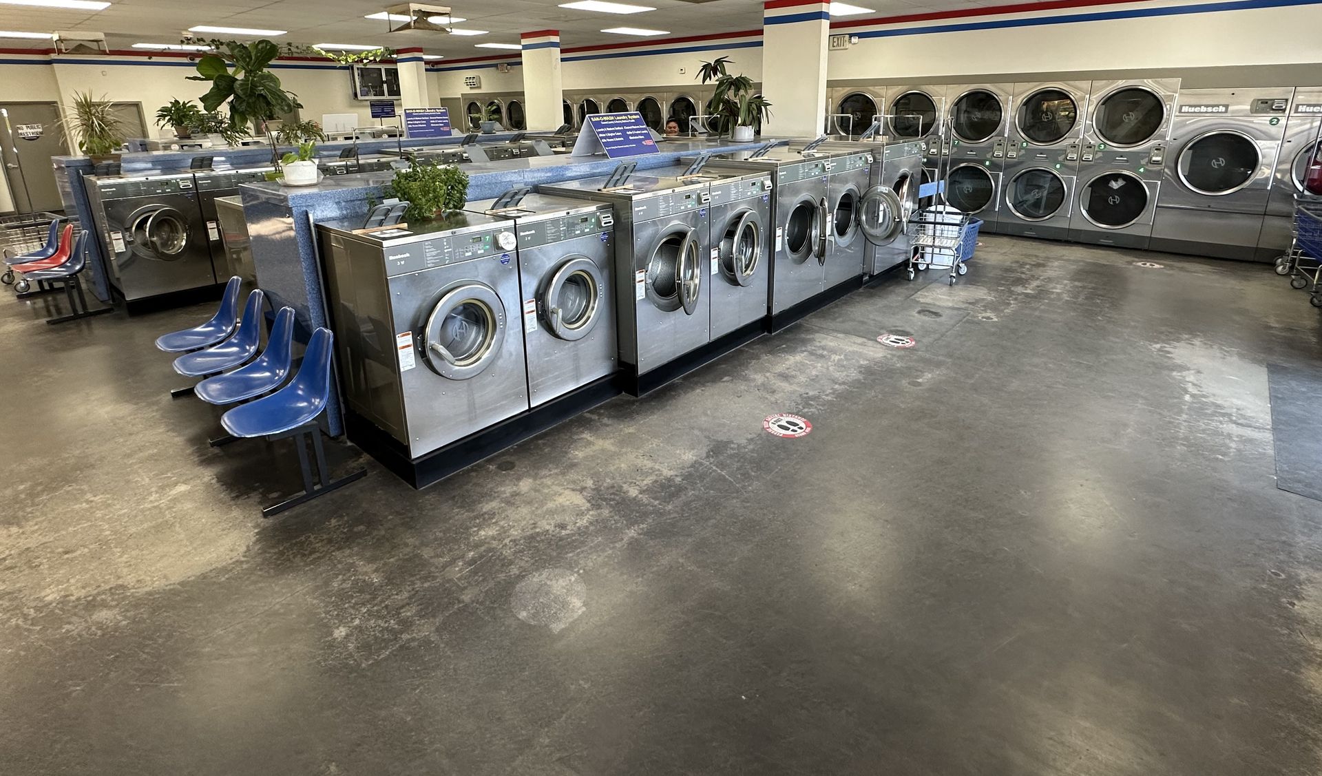 A wide interior view with a red-white-and-blue ceiling stripe, molded blue bucket seating along a row of washers, and a 'SAN-O-WASH Laundry System' sign.
