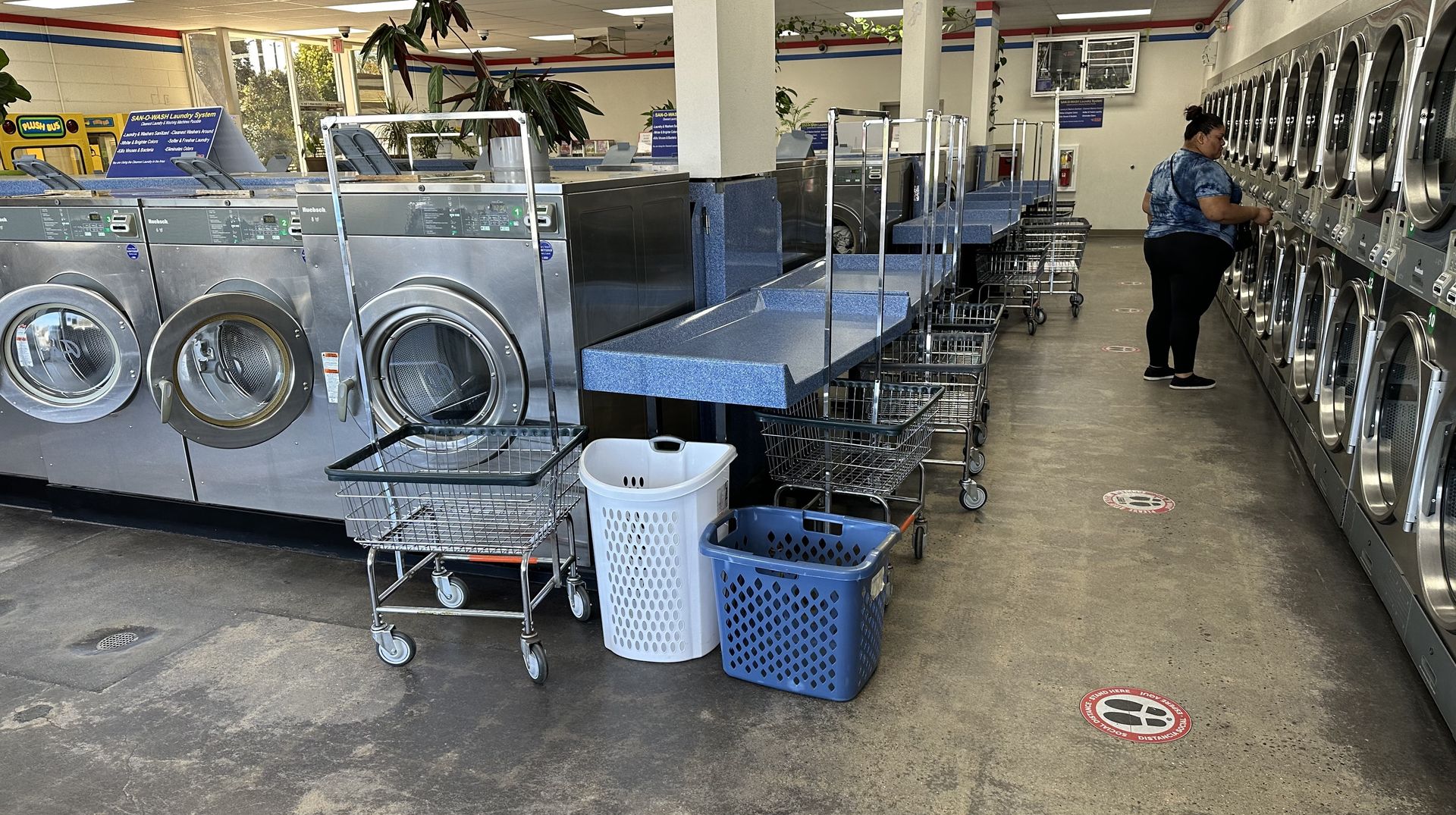 Rolling wire laundry carts and plastic baskets beside a speckled-blue folding counter, with a customer loading a washer along the far wall.