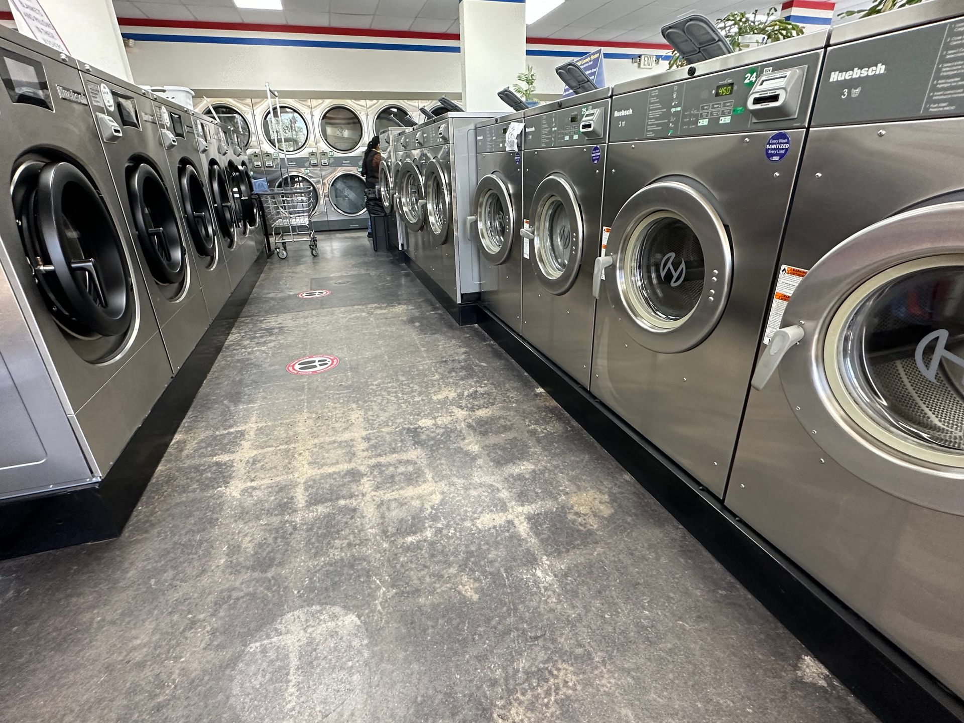 A center aisle between two rows of front-load washers marked 'Every Wash Sanitized', with a customer at the machines and a rolling cart in the foreground.