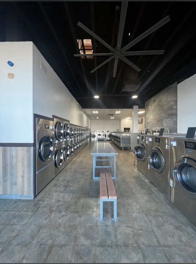 A laundromat aisle beneath a black slat ceiling with a large paddle fan, stacked dryers on one side and front-load washers on the other, with a bench down the center.