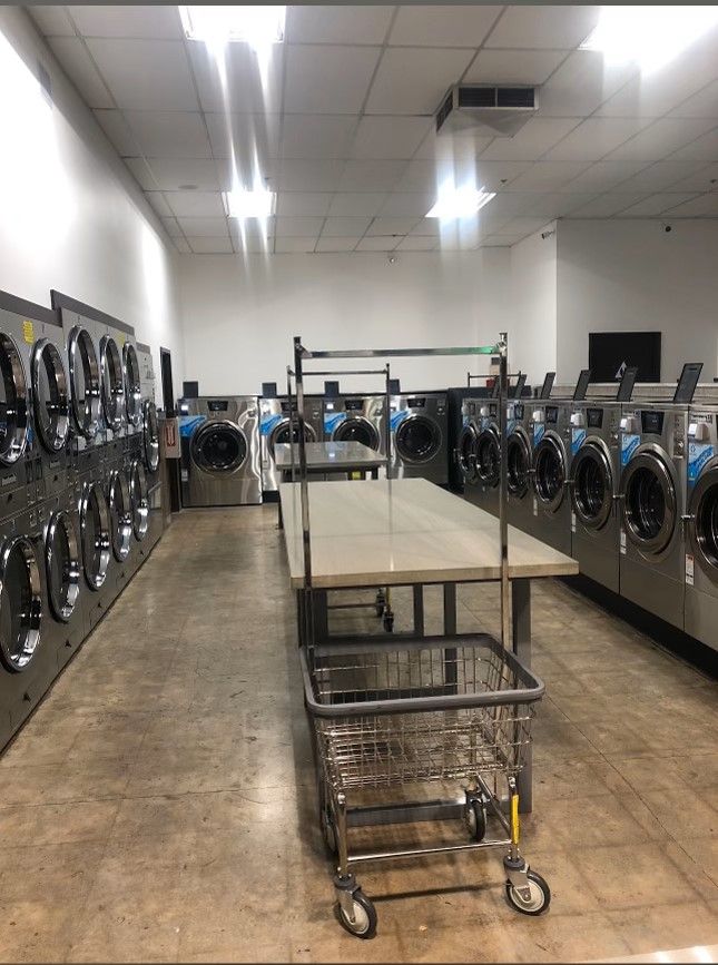 A laundromat interior with a wall of stacked dryers, two facing rows of front-load washers, a rolling wire cart, and a marble-look folding table in the foreground.