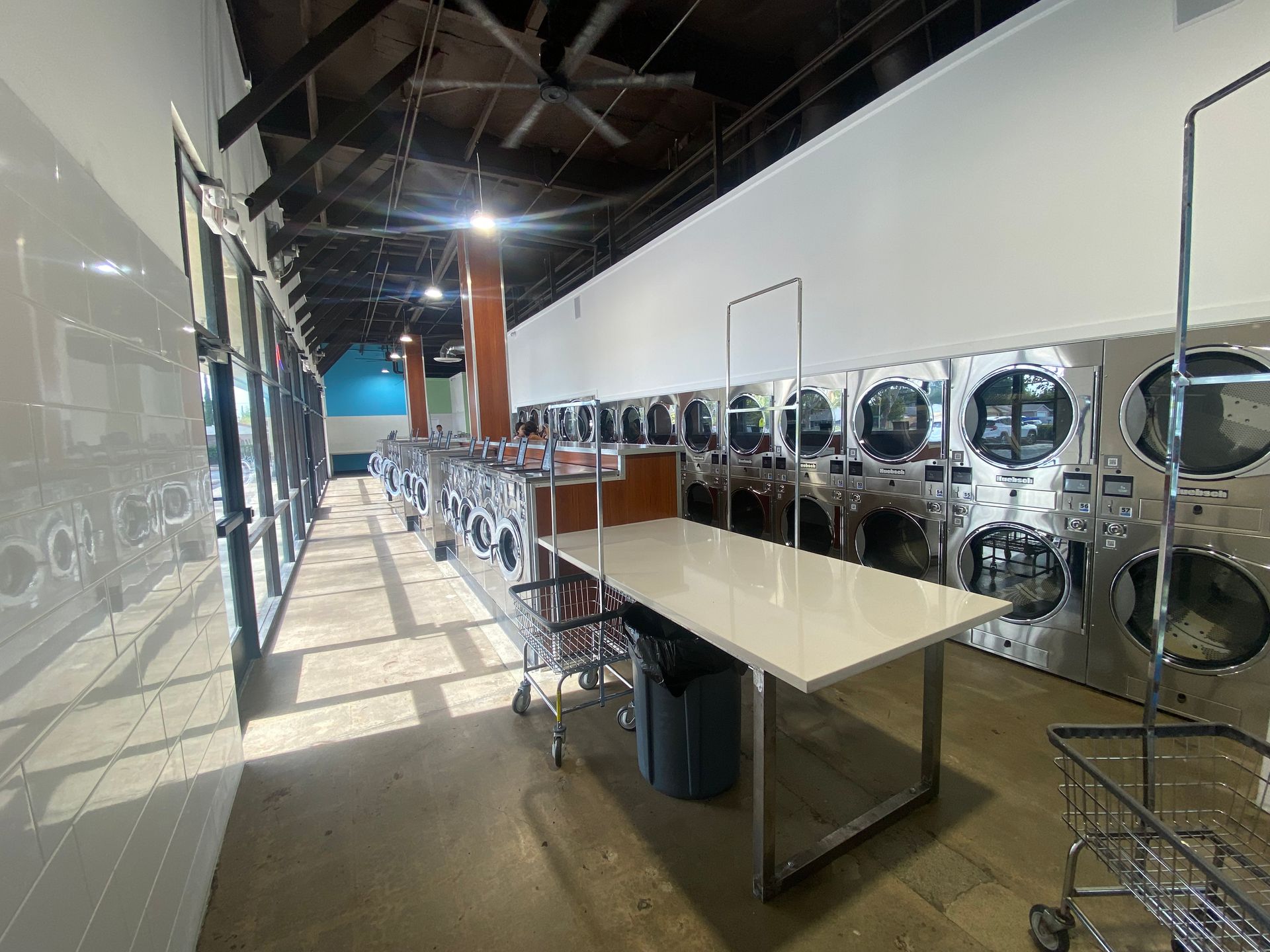 An interior aisle beneath exposed dark roof trusses and an industrial paddle fan, with a glass storefront on one side and a row of washers with a wood-topped counter on the other.